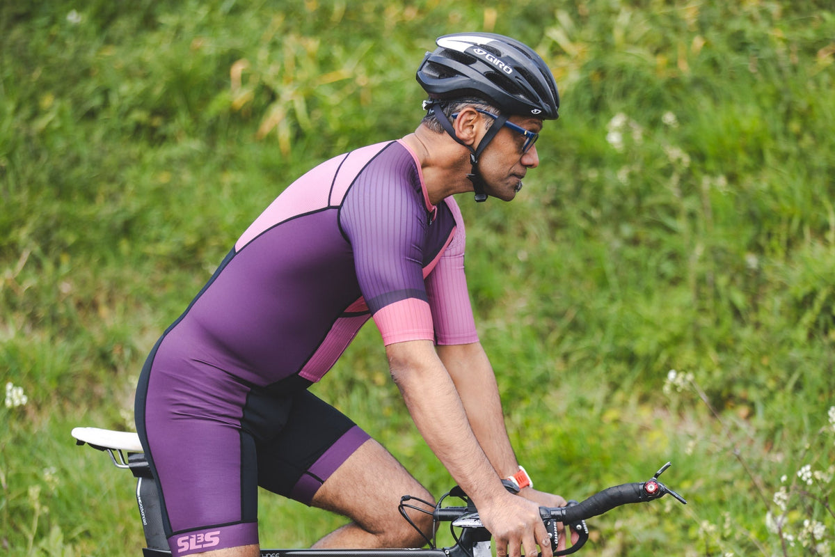 Cyclist wearing a purple and pink SLS3 triathlon suit, helmet, and sunglasses riding a road bike outdoors on a green, tree-lined road.
