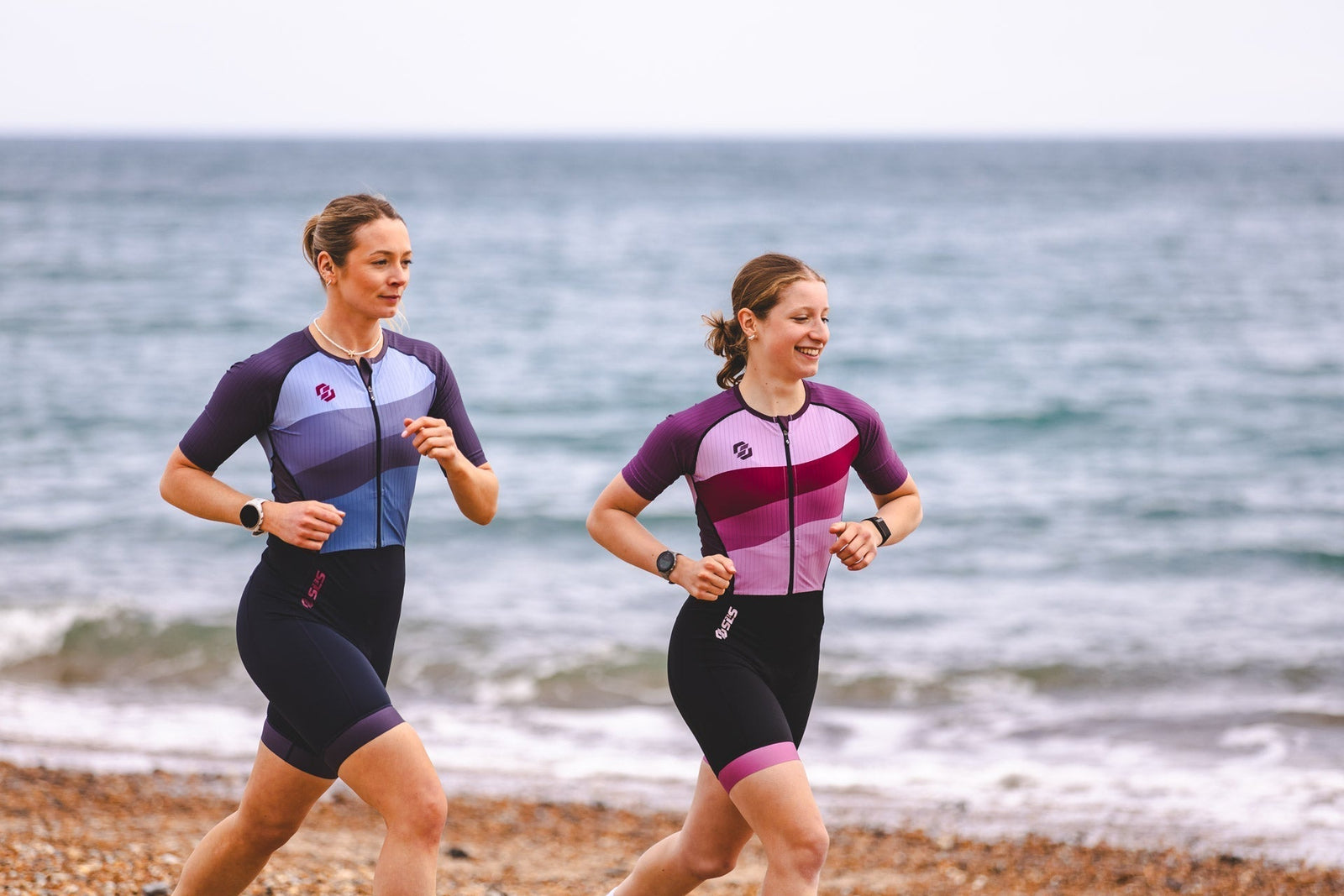 Two female triathletes running along the shoreline wearing SLS3 one-piece triathlon suits during an outdoor training session, showcasing women's performance tri suits designed for comfort and race-day performance.