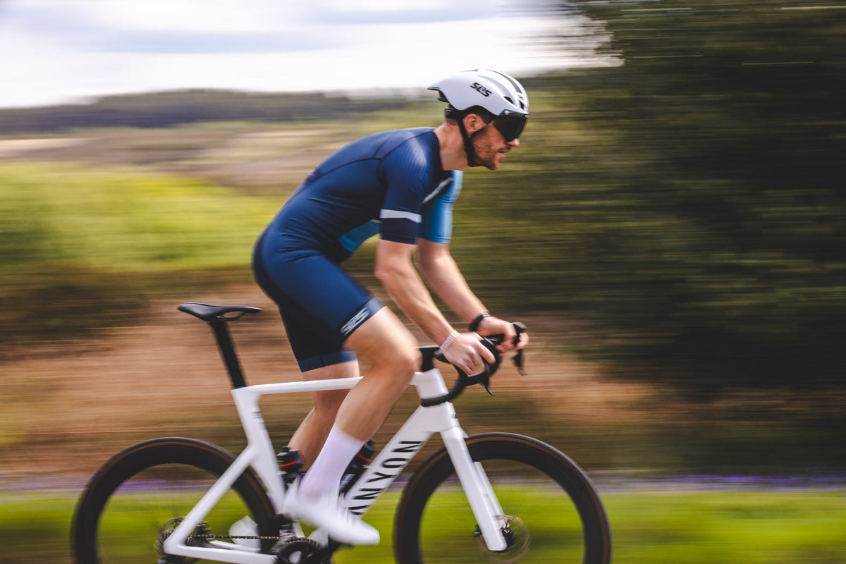 Male triathlete riding a time trial bike in an aero position on a rural road, wearing a navy SLS3 tri suit, cycling helmet, and sunglasses.