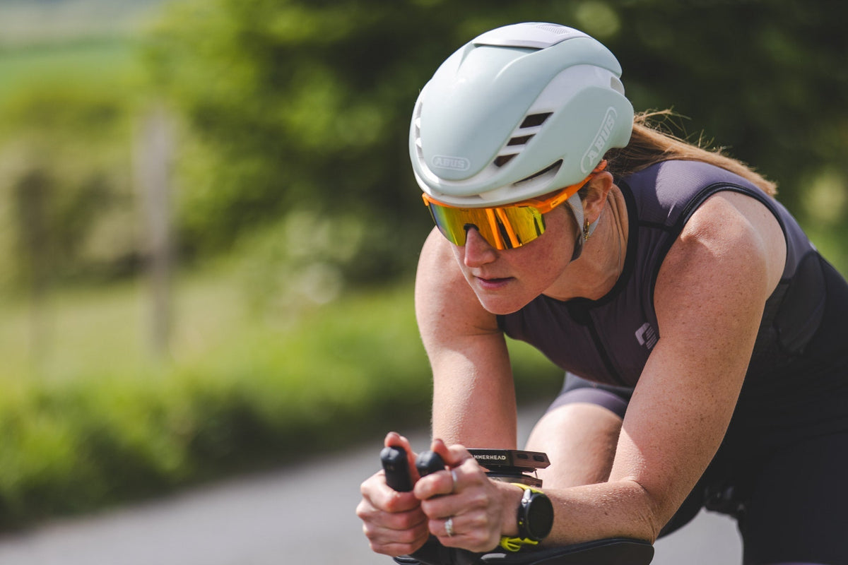 Female cyclist wearing a white helmet and sunglasses riding a road bike outdoors on a sunny, tree-lined road.