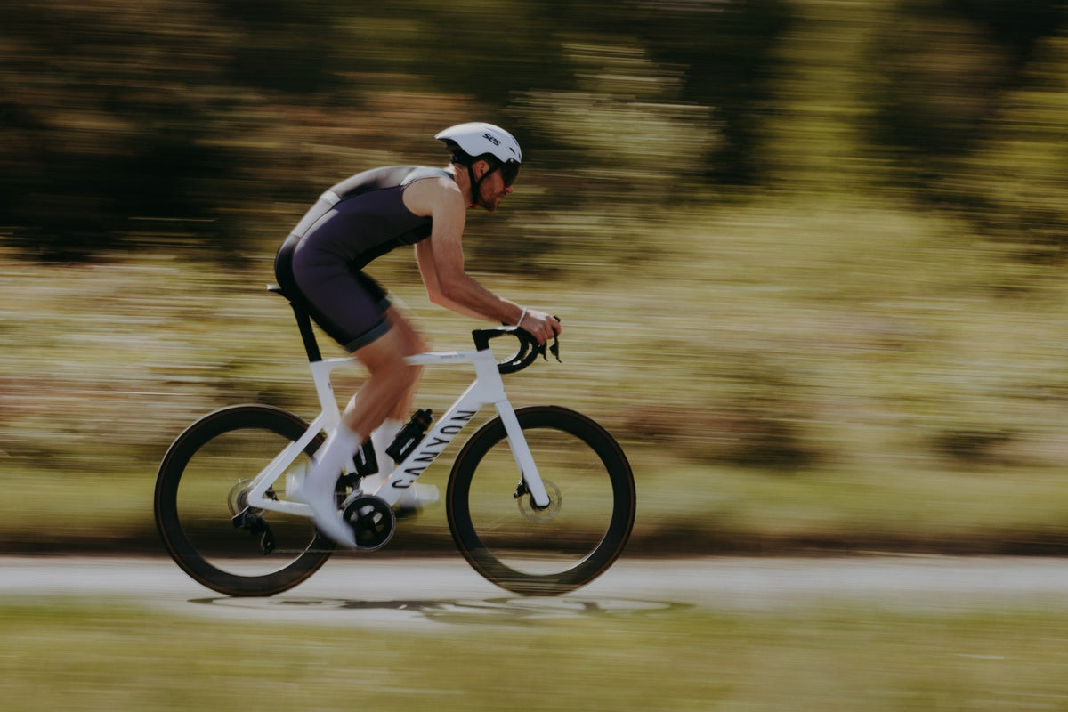 Male triathlete cycling at speed on a road bike while wearing a one-piece triathlon suit, with motion blur highlighting fast-paced outdoor training and race-day performance.